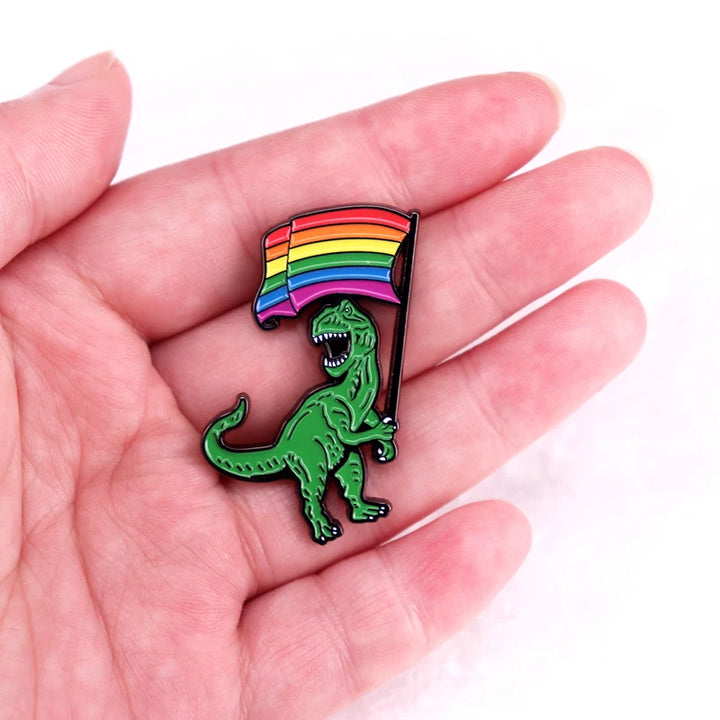 Ein metallischer Ansteck-Pin mit der Darstellung eines gruenen Dinosauriers (Tyrannosaurus Rex), der eine bunte Regenbogenfahne hochhaelt. Die Flagge zeigt lebendige Farben, die Vielfalt und Inklusion symbolisieren. Der Dinosaurier ist detailliert gestaltet und wirkt dynamisch, mit offenem Maul in einer kraftvollen Pose.