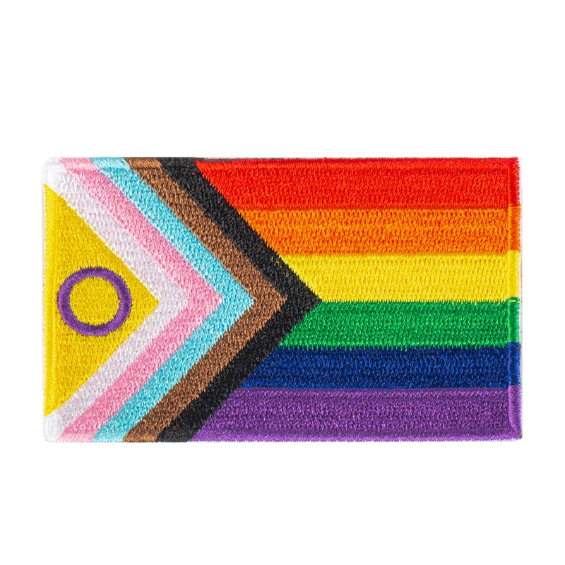 Aufnaeher mit der „Progress Pride Flagge“. Die Flagge kombiniert die klassischen Regenbogenfarben mit zusaetzlichen Streifen in Schwarz, Braun, Hellblau, Rosa und Weiß, die als Pfeil am linken Rand der Flagge integriert sind. Diese zusaetzlichen Farben repraesentieren die LGBTQI+ Communities von People of Color, die Transgender-Community und Inter Menschen, die mit HIV/AIDS leben oder verstorben sind.