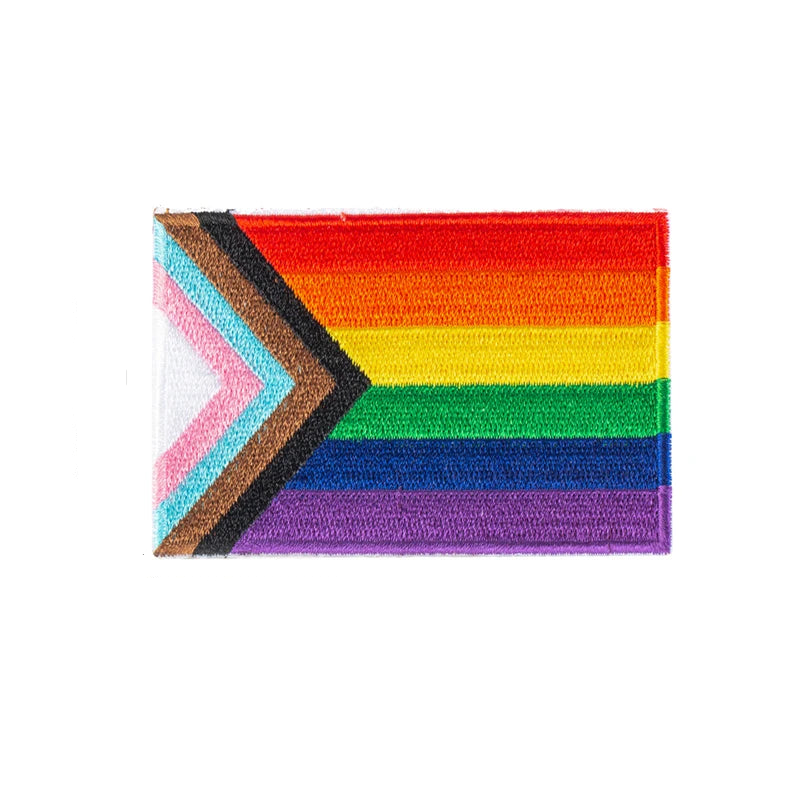 Aufnaeher mit der „Progress Pride Flagge“. Die Flagge kombiniert die klassischen Regenbogenfarben mit zusaetzlichen Streifen in Schwarz, Braun, Hellblau, Rosa und Weiß, die als Pfeil am linken Rand der Flagge integriert sind. Diese zusaetzlichen Farben repraesentieren die LGBTQ+ Communities von People of Color, die Transgender-Community und Menschen, die mit HIV/AIDS leben oder verstorben sind. Der Aufnaeher ist zum Aufnaehen gedacht und hat eine rechteckige Form.