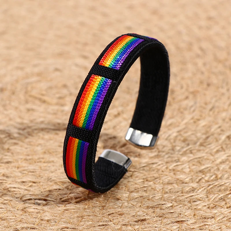 Ein schwarzer Armreif mit einem Regenbogenstreifen-Design in der Mitte, präsentiert auf einem strukturierten beigen Untergrund. Der Armreif hat silberne Endkappen und ein schlichtes, elegantes Design.