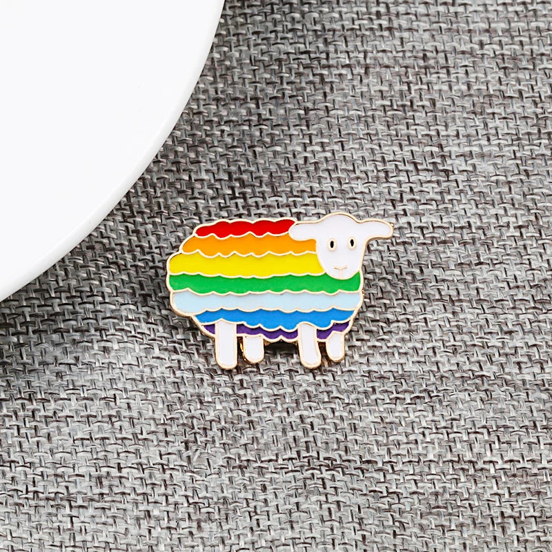 Ein Ansteck-Pin in Form eines Schafs mit einem Körper in den Farben der Regenbogenflagge. Der Pin liegt auf einem grauen Stoffhintergrund.