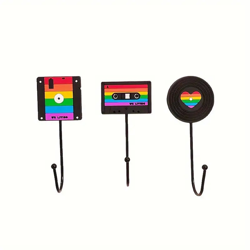 Die drei Wandhaken sind im Retro-Stil gestaltet: einer in Form einer Diskette, einer als Kassette und einer als Schallplatte. Jeder Haken ist bunt in Regenbogenfarben bemalt, was Stolz und Vielfalt symbolisiert.