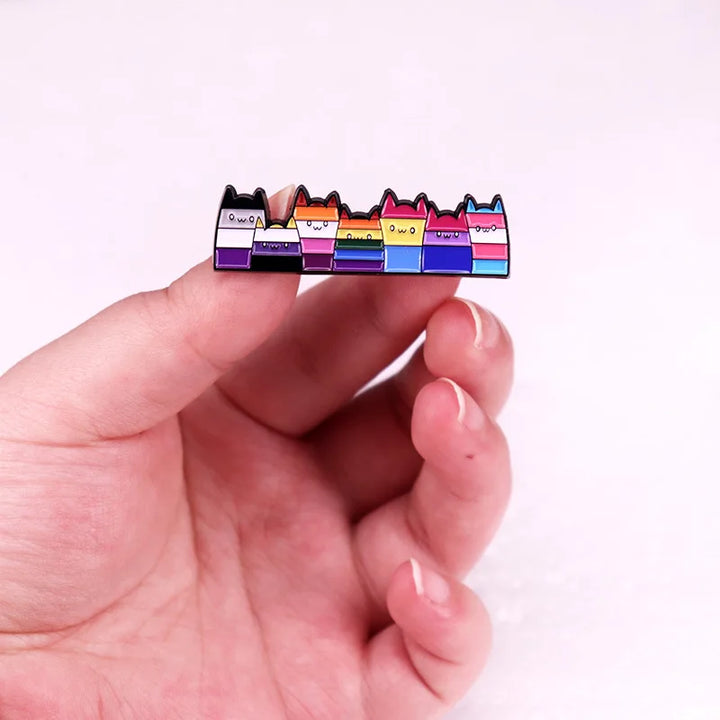 Reihe von Ansteckpins in Form kleiner Katzenfiguren, die jeweils die Farben verschiedener LGBTQ-plus -Pride-Flaggen tragen, darunter Lesben-, Bisexuellen-, Pansexuellen-, Nicht-binaeren-, Asexuellen- und Transgender-Flaggen. Die Katzen haben niedliche Gesichter und spitze Ohren, wodurch sie ein froehliches, verspieltes Aussehen erhalten.