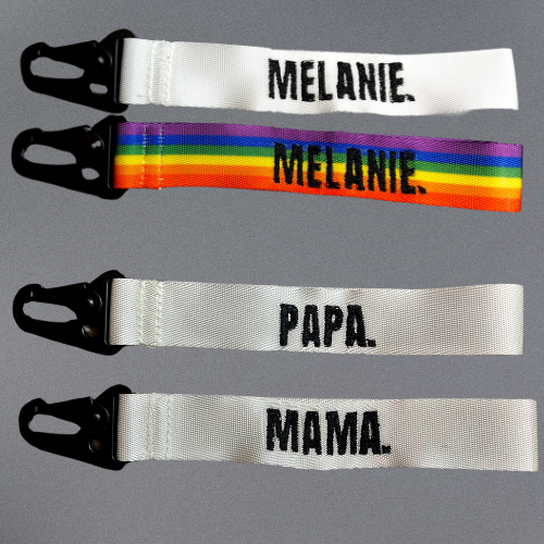 Vier Schlüsselanhänger aus Stoff mit Karabinerverschluss. Der obere ist weiß mit dem Schriftzug "MELANIE.", der zweite hat ein Regenbogenmuster mit "MELANIE.", der dritte ist weiß mit "PAPA.", und der vierte ist weiß mit "MAMA."