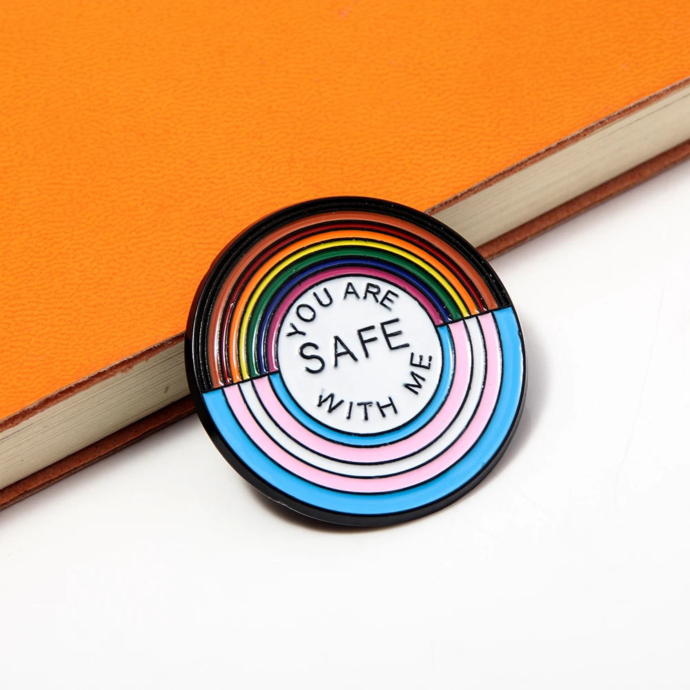 Ein runder Ansteck-Pin mit einem Design aus Regenbogenfarben und den Farben der Transgender-Flagge. In der Mitte steht der Text "YOU ARE SAFE WITH ME".