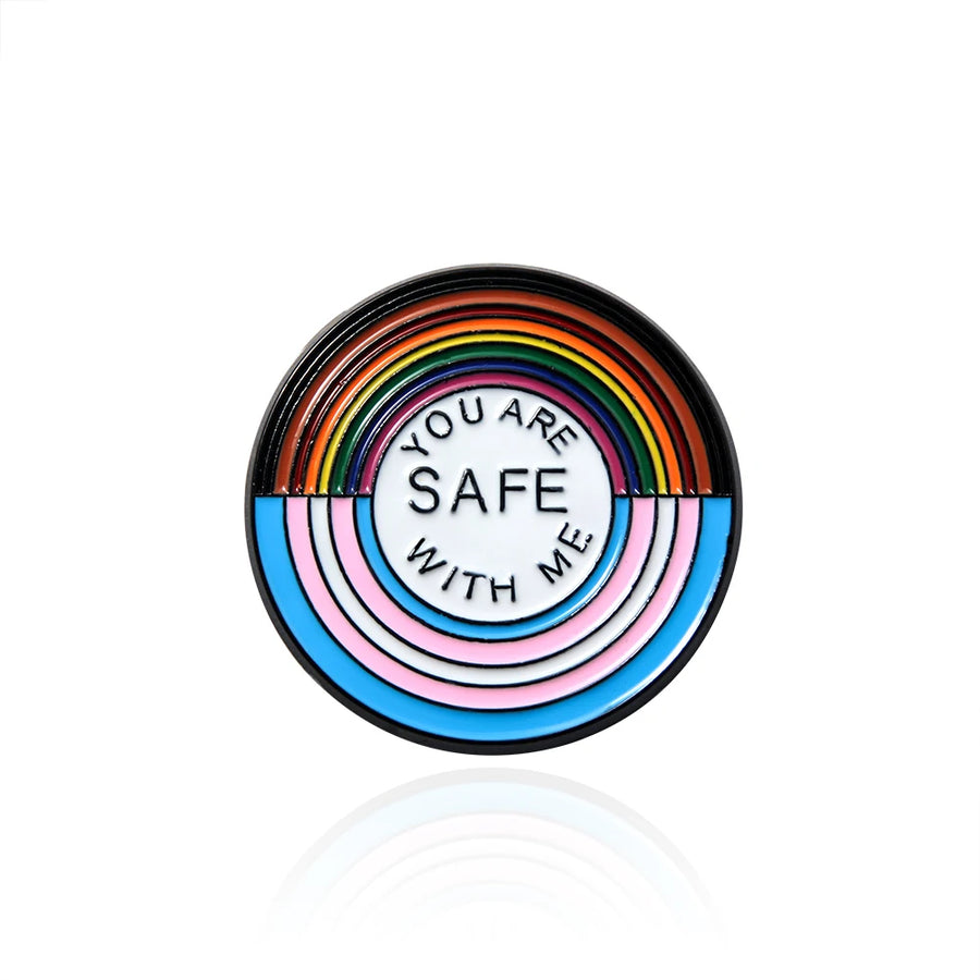 Ein runder Ansteck-Pin mit einem Design aus Regenbogenfarben und den Farben der Transgender-Flagge. In der Mitte steht der Text "YOU ARE SAFE WITH ME".